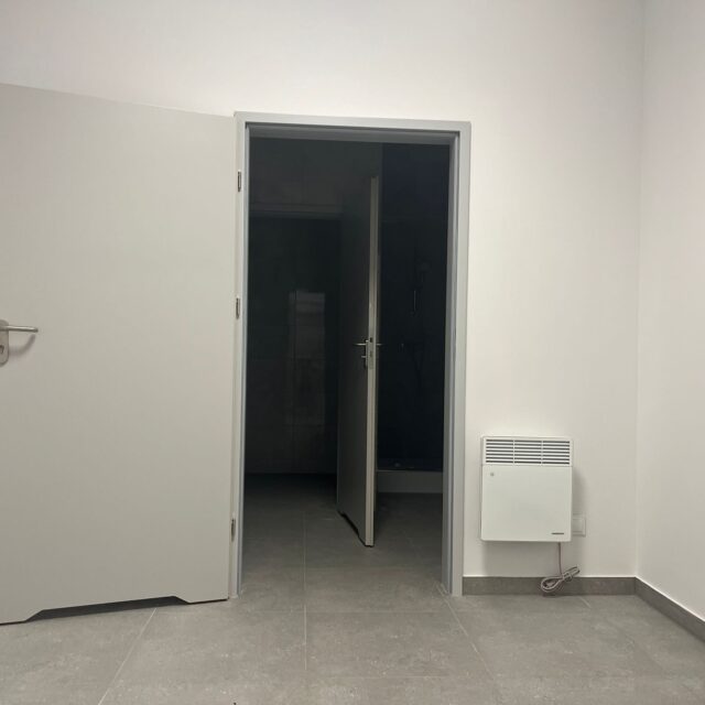 Widok wejścia do pomieszczeń sanitarnych z drzwiami prowadzącymi do strefy prysznicowej.