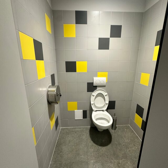 Kabina WC z podwieszaną miską ustępową i szarymi płytkami z żółtymi oraz czarnymi akcentami.