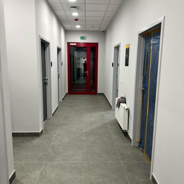 Korytarz z czerwonymi przeszklonymi drzwiami na końcu, szarymi drzwiami bocznymi i grzejnikiem.