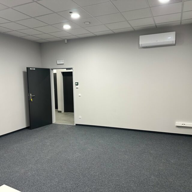 Pomieszczenie biurowe z ciemną wykładziną, klimatyzatorem ściennym i oświetleniem punktowym w suficie.