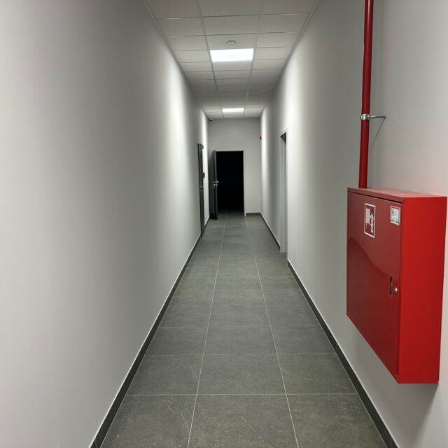 Długi korytarz z czerwoną szafką hydrantową i przewodem instalacji przeciwpożarowej na prawej ścianie.