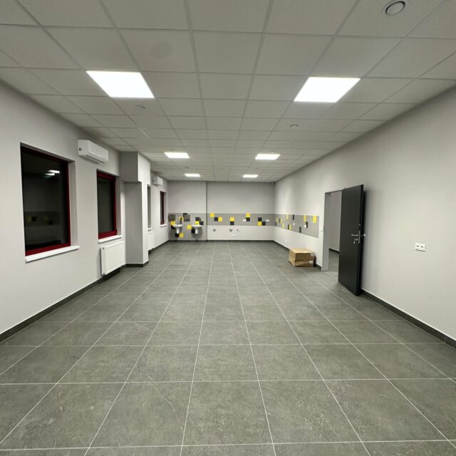 Sala z szarą posadzką, oknami i klimatyzatorami po lewej stronie oraz strefą umywalek w głębi.