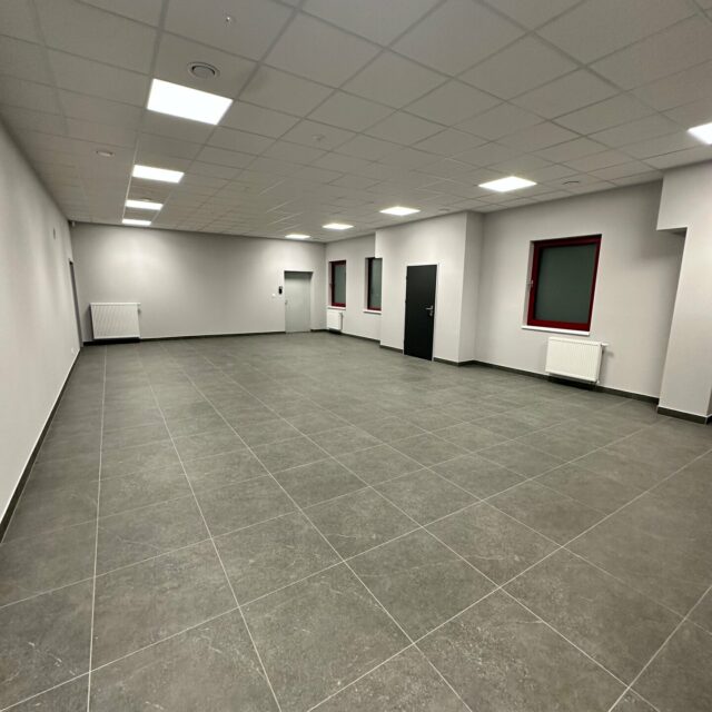 Przestronna sala z sufitem podwieszanym, panelami oświetleniowymi oraz oknami i grzejnikami po prawej stronie.