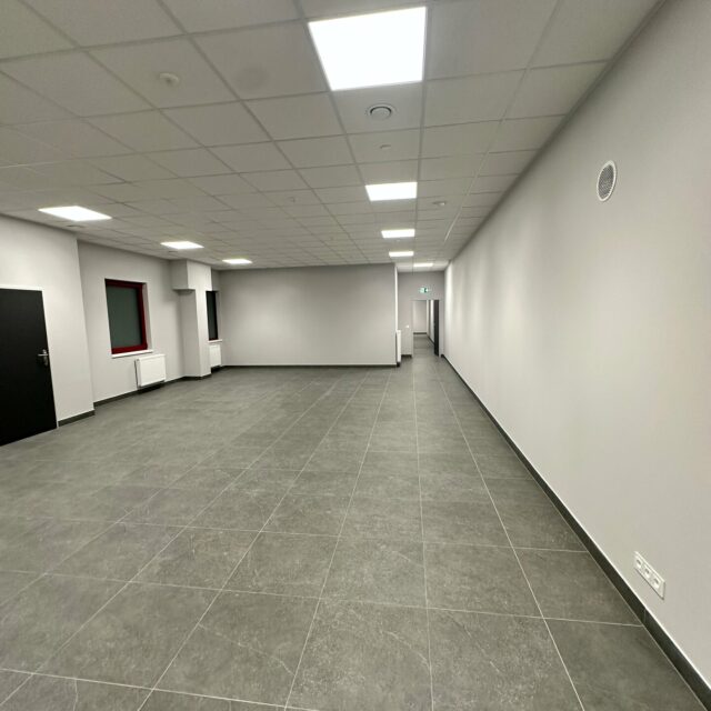 Widok dużej sali z jasnymi ścianami i szarą posadzką; po lewej stronie drzwi oraz okna z grzejnikami.