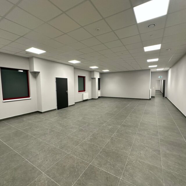 Sala z szarą posadzką z dużych płytek, oknami i grzejnikami wzdłuż lewej ściany oraz drzwiami w głębi.