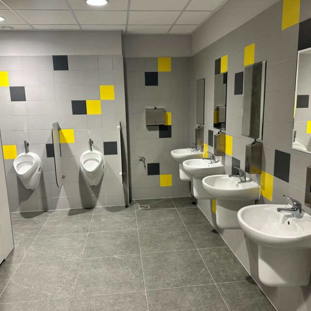 Dwa pisuary oraz trzy umywalki z lustrami w sanitariatach wykończonych szarymi, żółtymi i czarnymi płytkami.