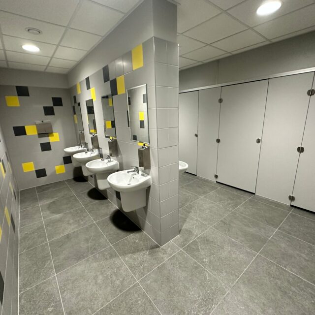 Sanitariaty z kabinami WC po prawej stronie, umywalkami przy ścianie działowej i ciemną posadzką z płytek.